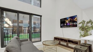 Un salon moderne avec un canapé gris, une table basse en verre, un téléviseur fixé au mur et une plante.