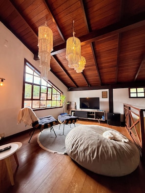 A spacious living room with a high ceiling, wooden beams, a large window, a television, and a bean bag. - Entire home in Florianópolis, Brazil near Lagoa, Mole and Joaquina (Lagoa da Conceição)