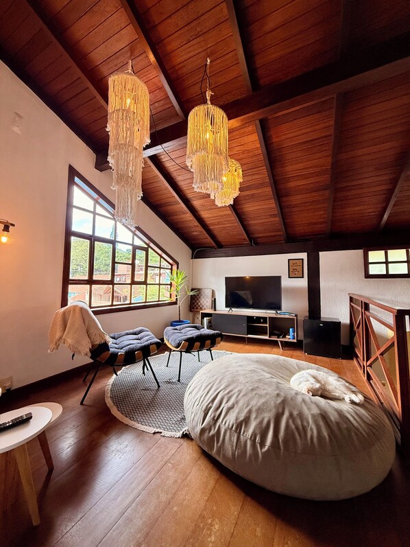Ein geräumiges Wohnzimmer mit hoher Decke, Holzbalken, einem großen Fenster, einem Fernseher und einem Sitzsack.