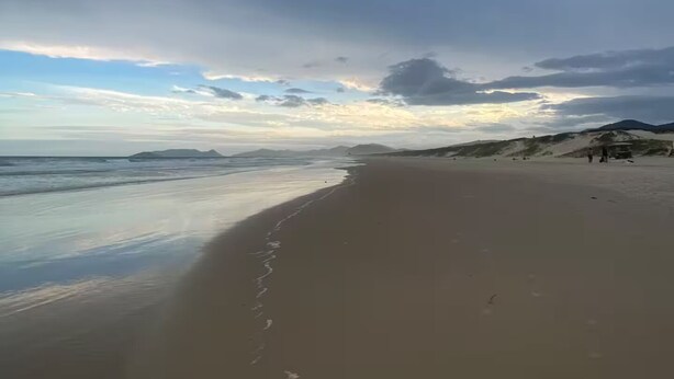 A beach with a wide sandy shore, a calm sea, and distant mountains under a cloudy sky. - Entire home in Florianópolis, Brazil near Lagoa, Mole and Joaquina (Lagoa da Conceição)