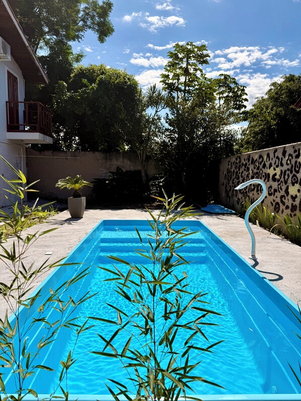 Une piscine rectangulaire à l’eau d’un bleu limpide, entourée de plantes et d’arbres, et un bâtiment avec un balcon en arrière-plan.