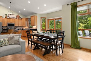 A dining area with a wooden table and chairs, a kitchen with wooden cabinets, and a view of greenery through large windows.