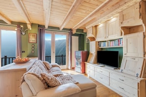 A cozy room with a sofa, a TV, wooden cabinets, and a view of mountains. - Baita Delle Stelle (Bessè Alto)