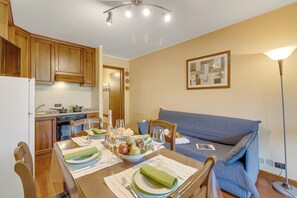 A dining area with a wooden table set for two, a blue sofa, and a kitchen in the background. - Appartamento Tracce Di Neve (Sestriere)