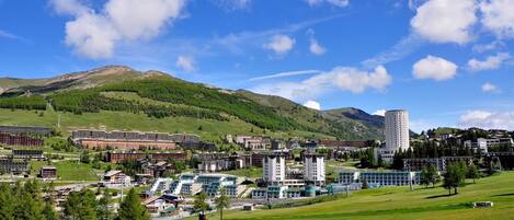 A town with modern buildings, a prominent tower, and a green hill in the background.