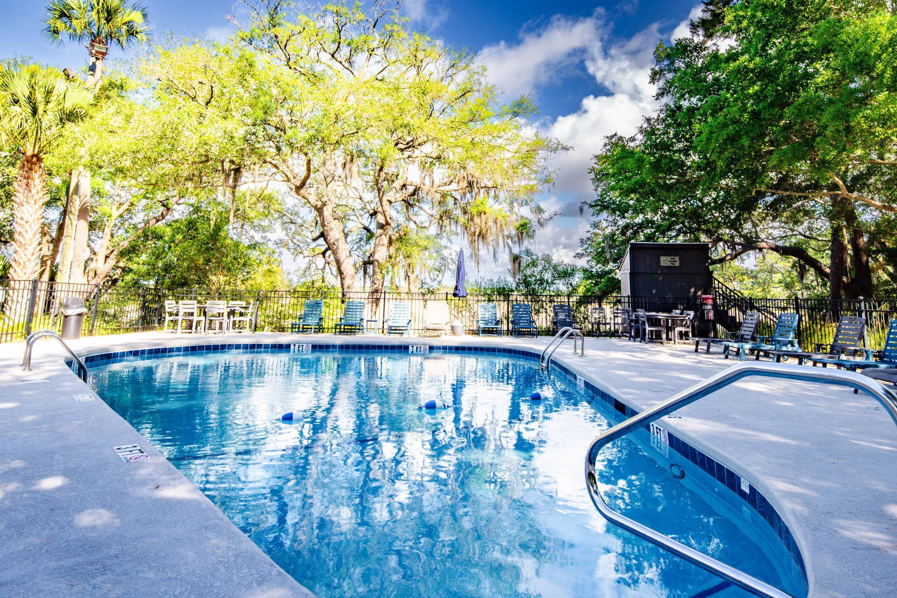 A swimming pool with lounge chairs and tables, surrounded by trees and a clear sky.