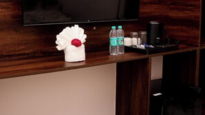 Ein Hotelzimmer mit einem an der Wand befestigten Flachbildfernseher, einem kleinen weißen Handtuch mit roter Blume, zwei Wasserflaschen, einer Kaffeemaschine und einem schwarzen Koffer auf einem Holzboden.