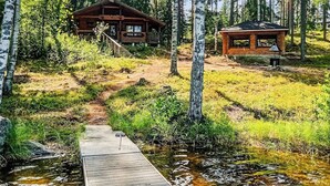 Uma cabana com um deck de madeira que dá para um pequeno cais sobre um riacho, cercada por uma floresta densa.