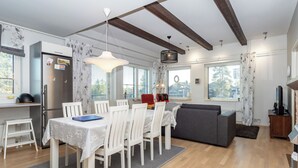 Eine moderne Küche mit Einbaukühlschrank, Essbereich mit weißen Stühlen und einem Tisch, ein graues Sofa und ein TV-Schrank.