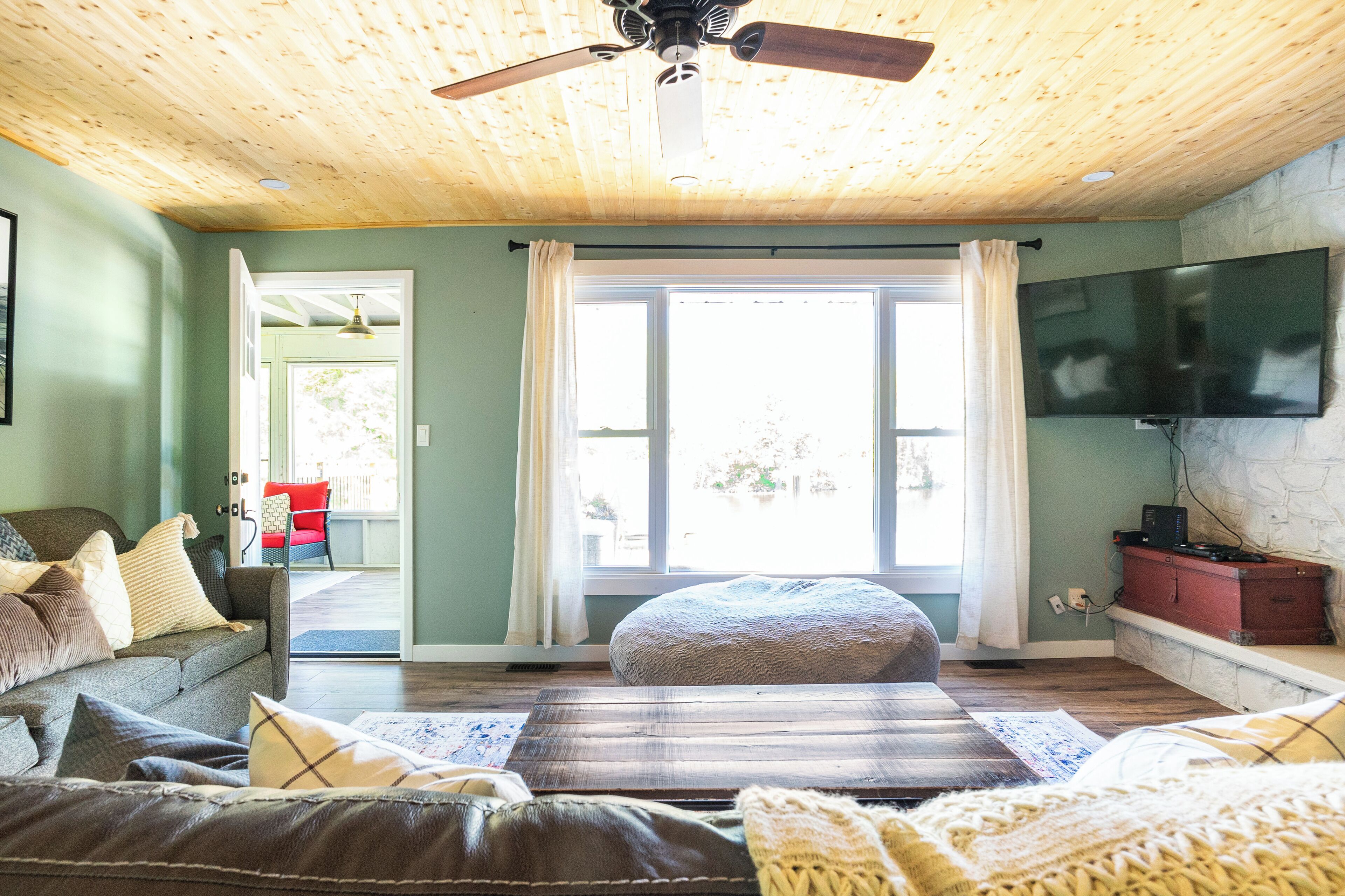 A cozy living room with a wooden ceiling, a large window with curtains, a flat-screen TV, and a comfortable seating area with a sofa and ottoman.