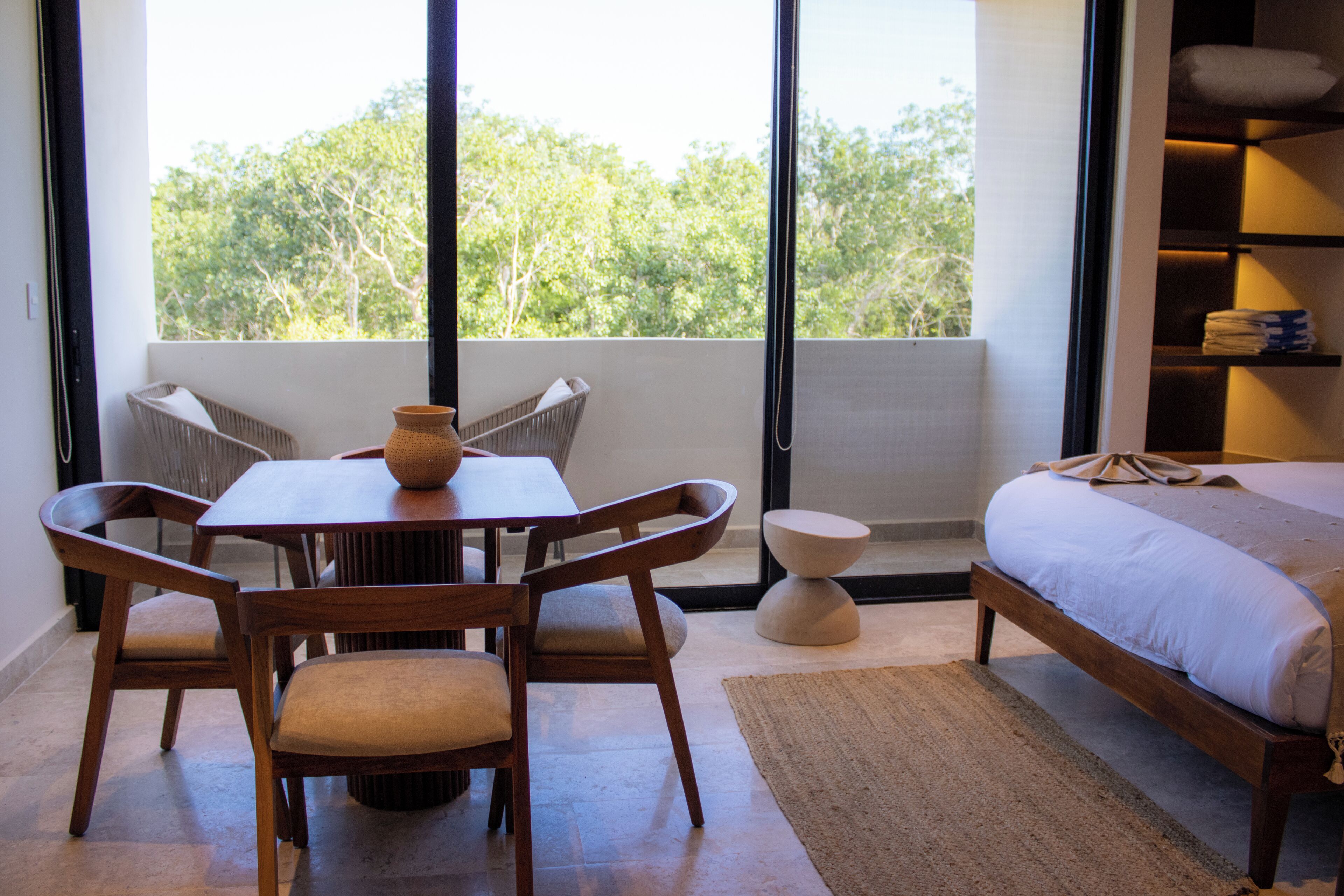 Una habitación con un ventanal grande, una mesa de comedor de madera con sillas, una mesita redonda pequeña, una cama con colchón blanco y un estante con toallas dobladas.