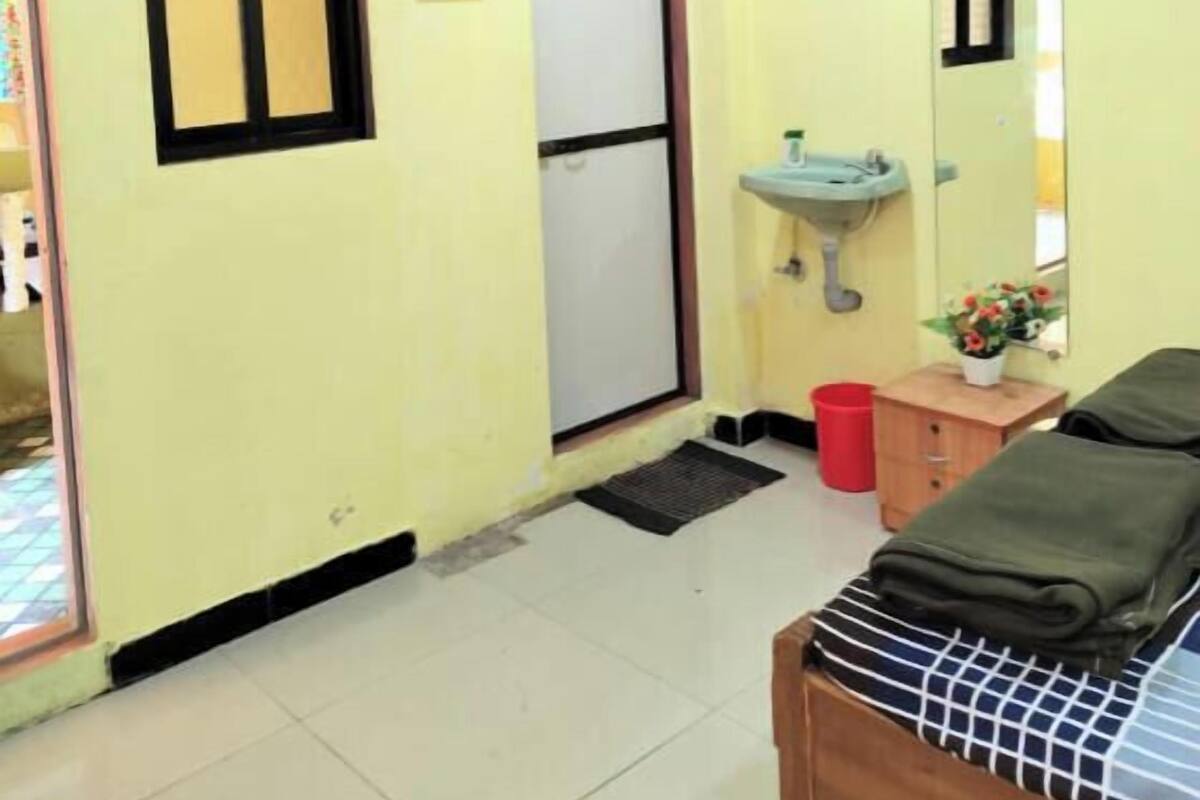 Une pièce avec un plafond en bois, un ventilateur de plafond, une fenêtre à cadre noir, une porte blanche, un lavabo avec un miroir, un petit meuble en bois et un lit recouvert d’une courtepointe à rayures.