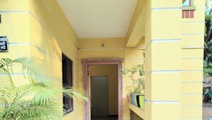 A narrow, yellow corridor with a tiled floor leading to a door. There are potted plants on the sides and a small step leading up to the entrance. - Mauli cottage (Murud)