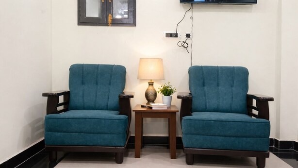 Two teal armchairs with wooden armrests, a small wooden side table with a lamp and a plant, and a wall-mounted TV. - Hotel Moon Year (Lucknow)