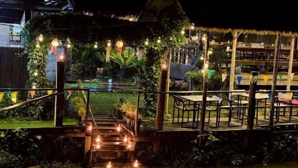 Ein Außenbereich bei Nacht mit einer Holzterrasse, Stufen, die zu einer kleinen Brücke über dem Wasser führen, und Lichterketten.
