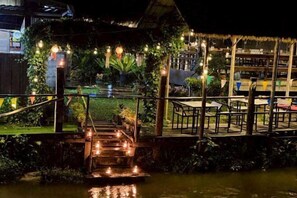 A nighttime outdoor area with a wooden deck, steps leading to a small bridge over water, and string lights. - Banna Cafe & Glamping (Mon Pin)