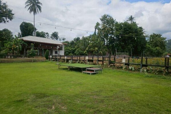 A grassy outdoor area with a wooden fence, a small building, and trees in the background. - Banna Cafe & Glamping (Mon Pin)