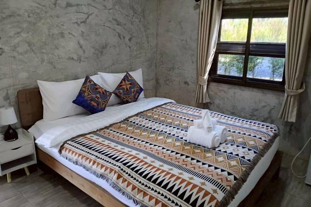 Ein Bett mit gemusterten Textilien, ein Nachttisch mit Lampe und ein Fenster mit Vorhängen.