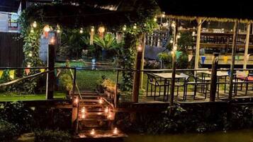 A nighttime outdoor area with a wooden bridge over water, surrounded by greenery and string lights.