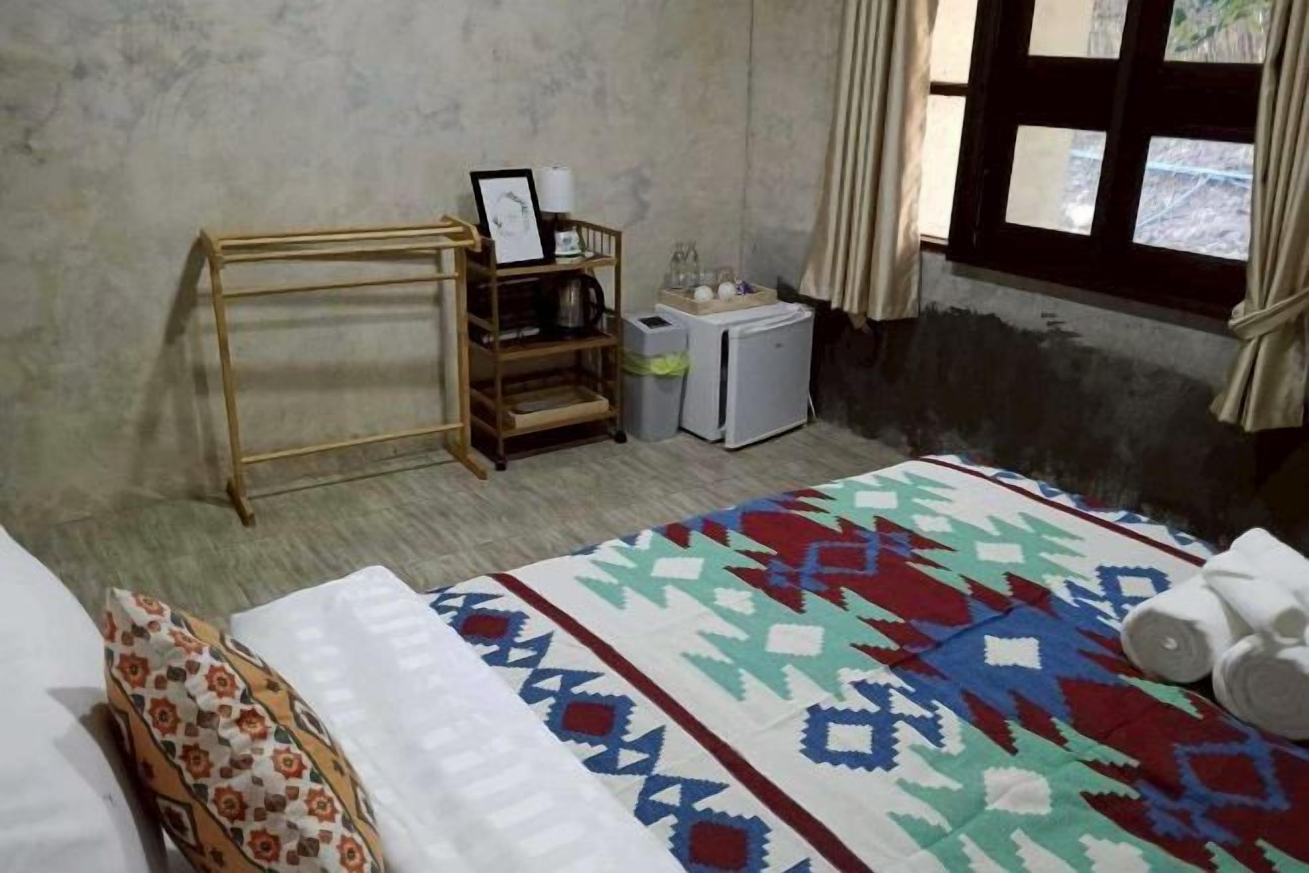 A bedroom with a bed, a wooden shelf with a picture frame, a small refrigerator, and a window with curtains.