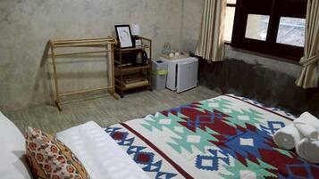 A bedroom with a bed, a wooden shelf with a picture frame, a small refrigerator, and a window with curtains.