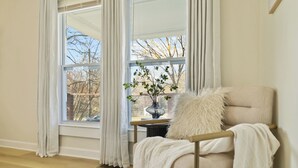 Habitación de esquina con un sillón color crema, una manta blanca, un florero con plantas verdes y vistas a árboles a través de amplias ventanas.