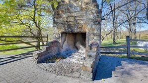 A stone fireplace with a fire, surrounded by a paved area and a wooden fence. - Walk to Golf Course: Spacious Rough River Retreat! (Falls of Rough)