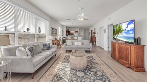 Ein geräumiges Wohnzimmer mit einem grauen Sofa, einem runden Hocker und einem großen Flachbildfernseher auf einer Holzkommode.