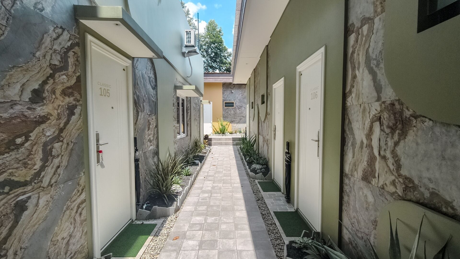 A narrow corridor with doors labeled 105 and 106, leading to a small outdoor area with plants and a tiled path.