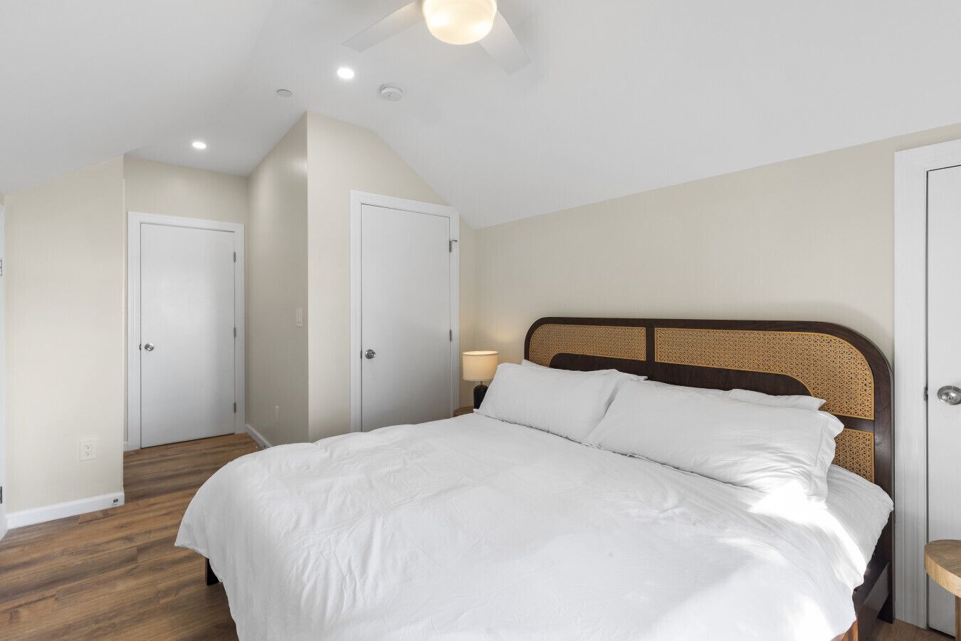 Quarto com uma cama grande, piso de madeira e paredes brancas. Há duas portas e uma luminária no teto.