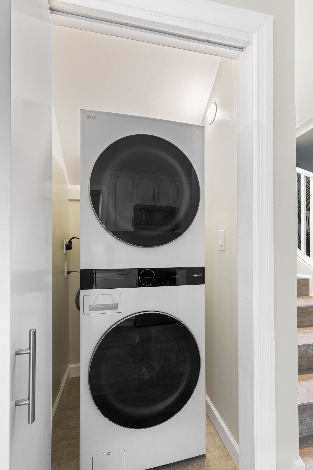 A compact, white, wall-mounted washer and dryer unit in a hallway with a wooden floor and a staircase in the background.
