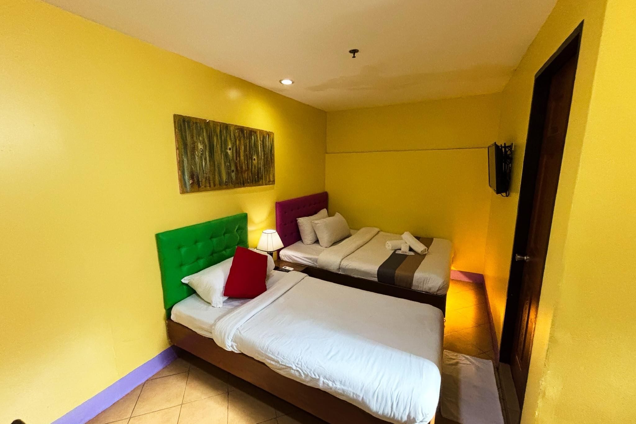 A room with two beds, one with a green headboard and the other with a purple headboard, both with white bedding. There is a wall-mounted artwork above the beds and a small table with a lamp between them.