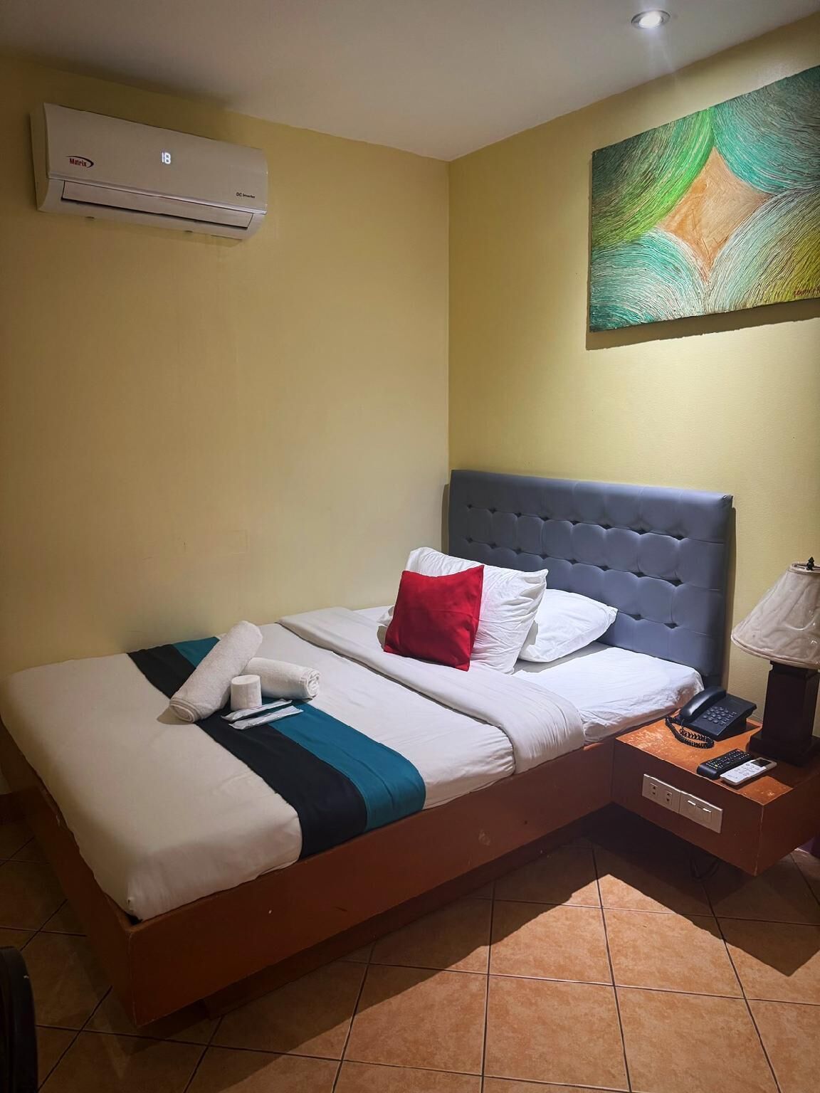 A hotel room with a bed, a nightstand, a lamp, an air conditioner, and a painting on the wall.