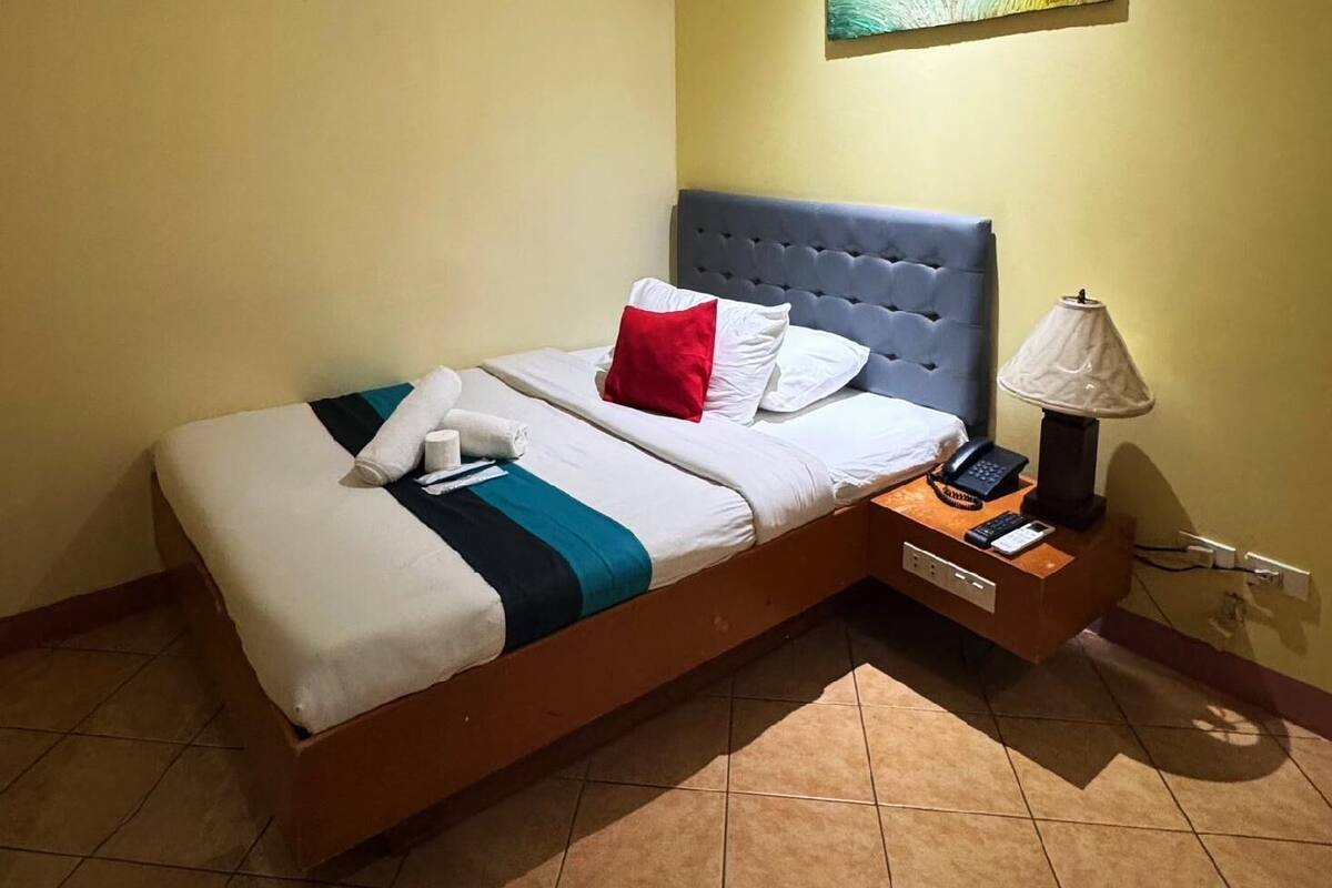 A hotel room with a bed, a bedside table with a lamp, an air conditioner, and a painting on the wall.