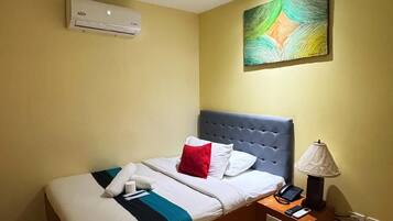 A hotel room with a bed, a bedside table with a lamp, an air conditioner, and a painting on the wall.