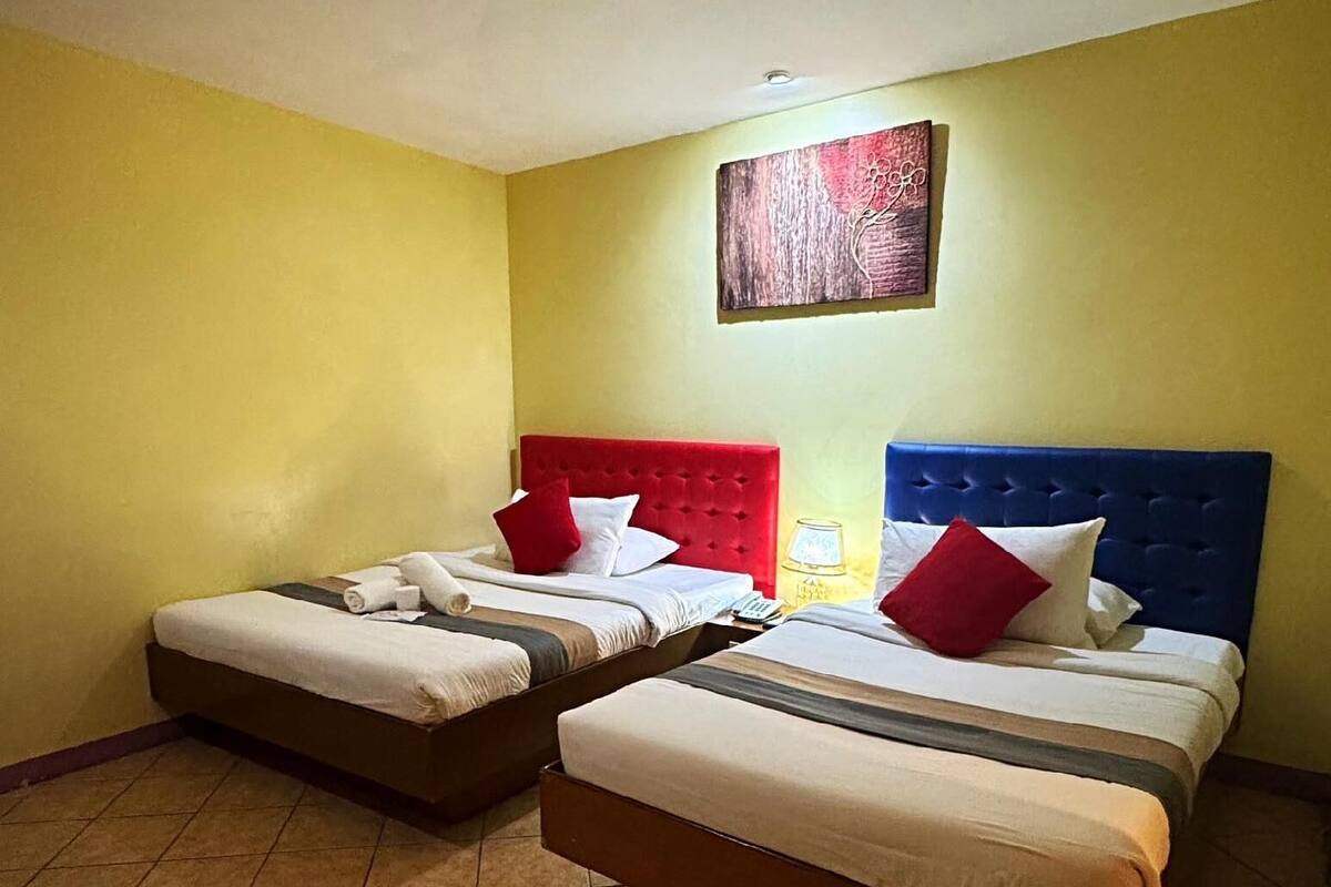 A hotel room with two beds, each with a different colored headboard, a painting on the wall, and a tiled floor.