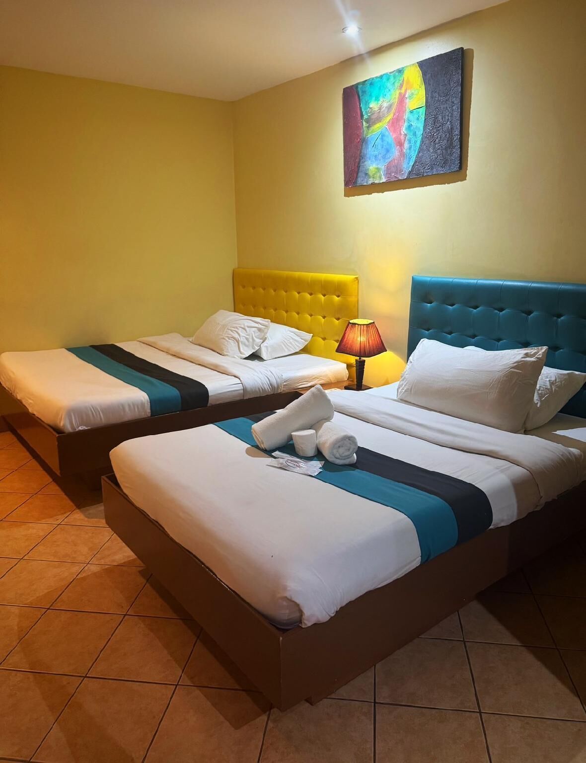 Habitación de hotel con dos camas, cada una con cabecero, un cuadro abstracto de colores en la pared y suelo de baldosas.