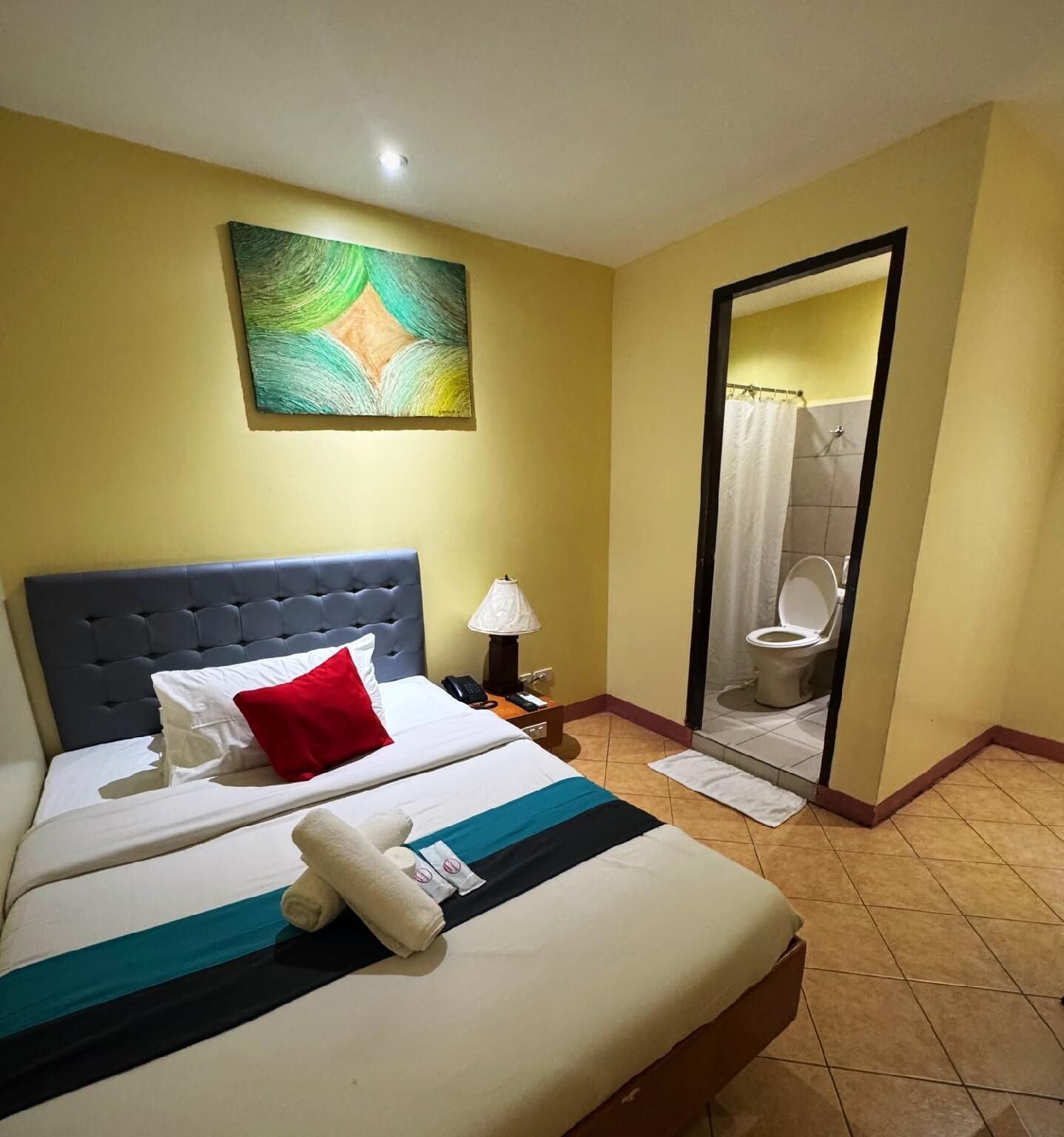 A hotel room with a bed, a nightstand, a lamp, a painting on the wall, and a bathroom visible through an open door.