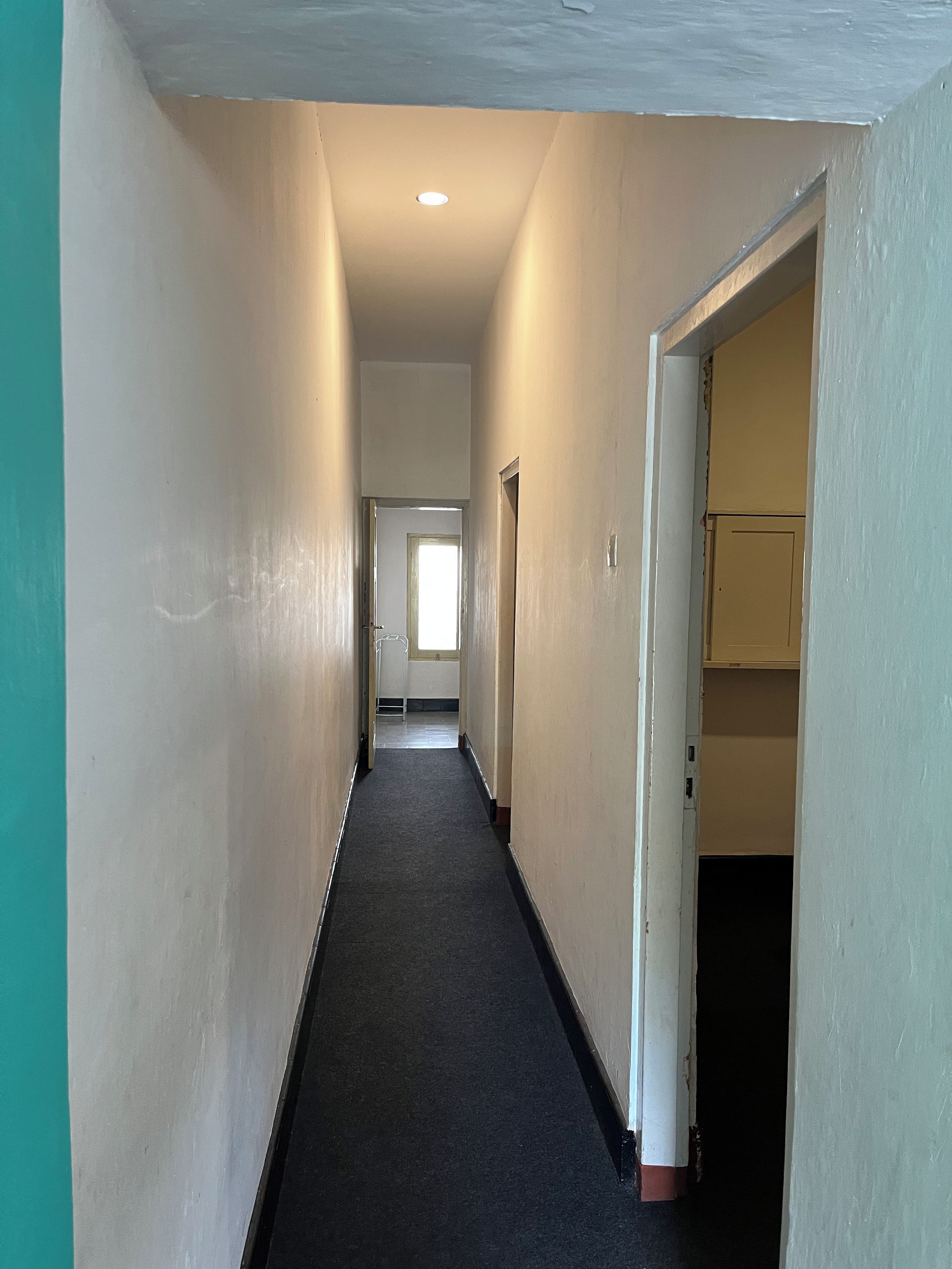 Un couloir étroit, dont le sol est recouvert d’une moquette, mène à une pièce dotée d’une porte et d’une fenêtre.