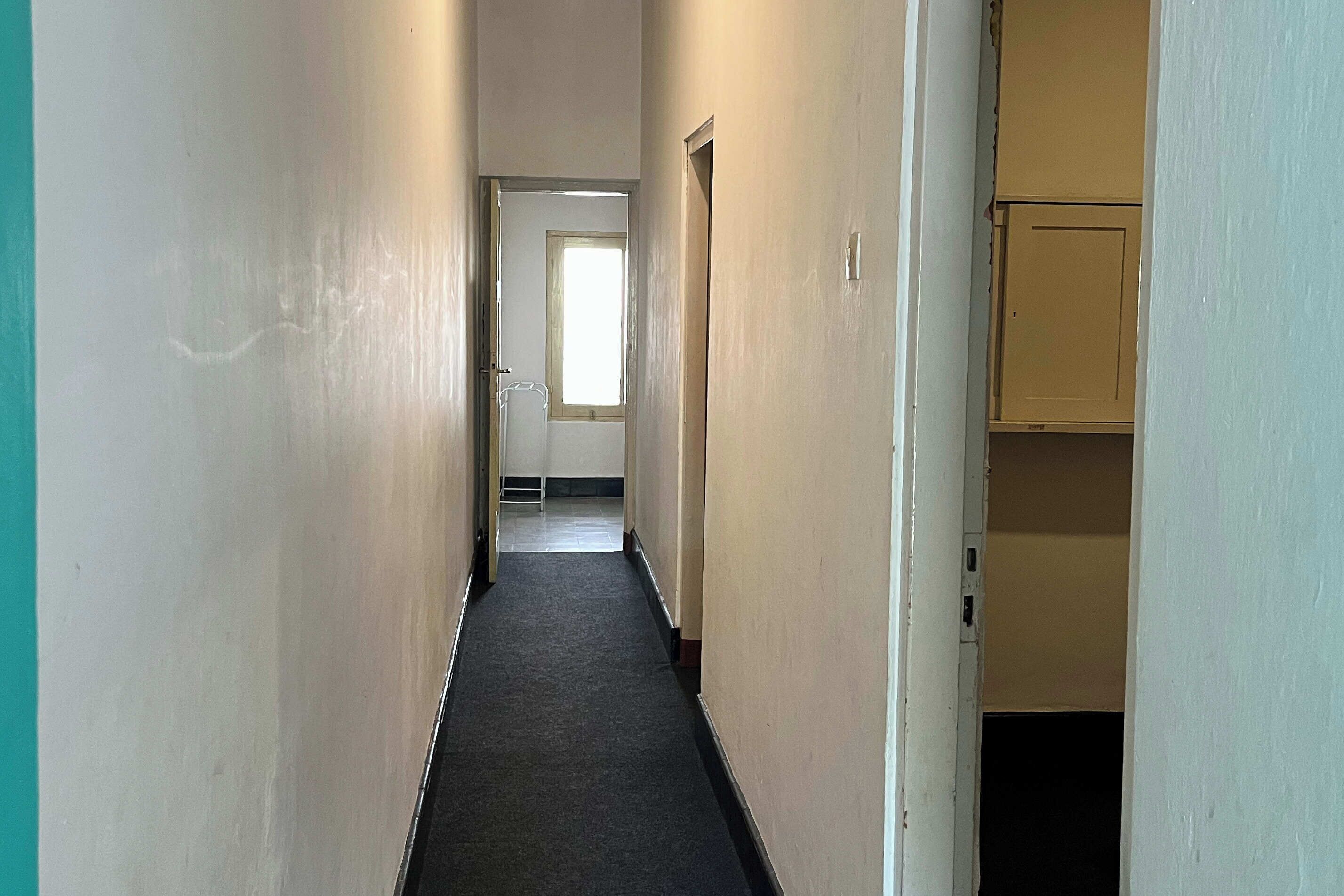 A narrow hallway with a carpeted floor, leading to a room with a door and a window.