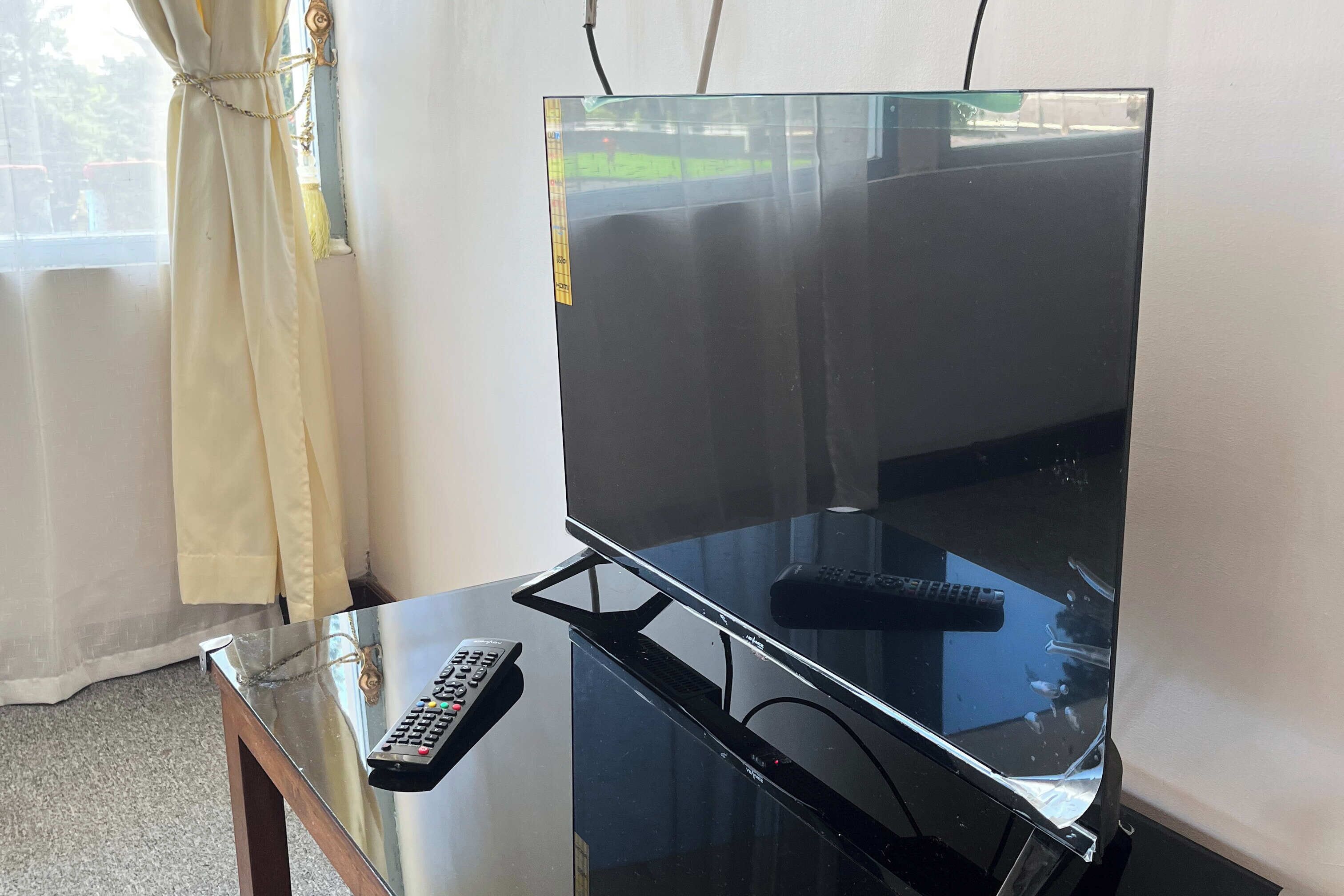 Un téléviseur à écran plat posé sur une table en verre, avec une télécommande à proximité, une fenêtre avec des rideaux et un reflet visible de l’extérieur.