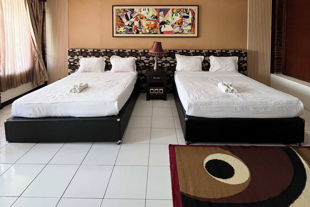 A hotel room with two beds, a nightstand, a lamp, a colorful abstract painting, and a patterned rug.
