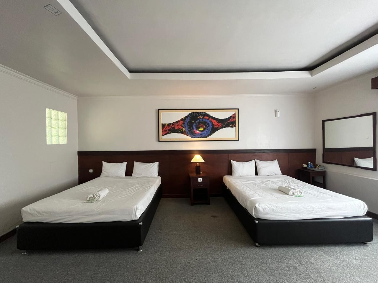 A hotel room with two beds, a nightstand with a lamp, a painting on the wall, and a mirror.