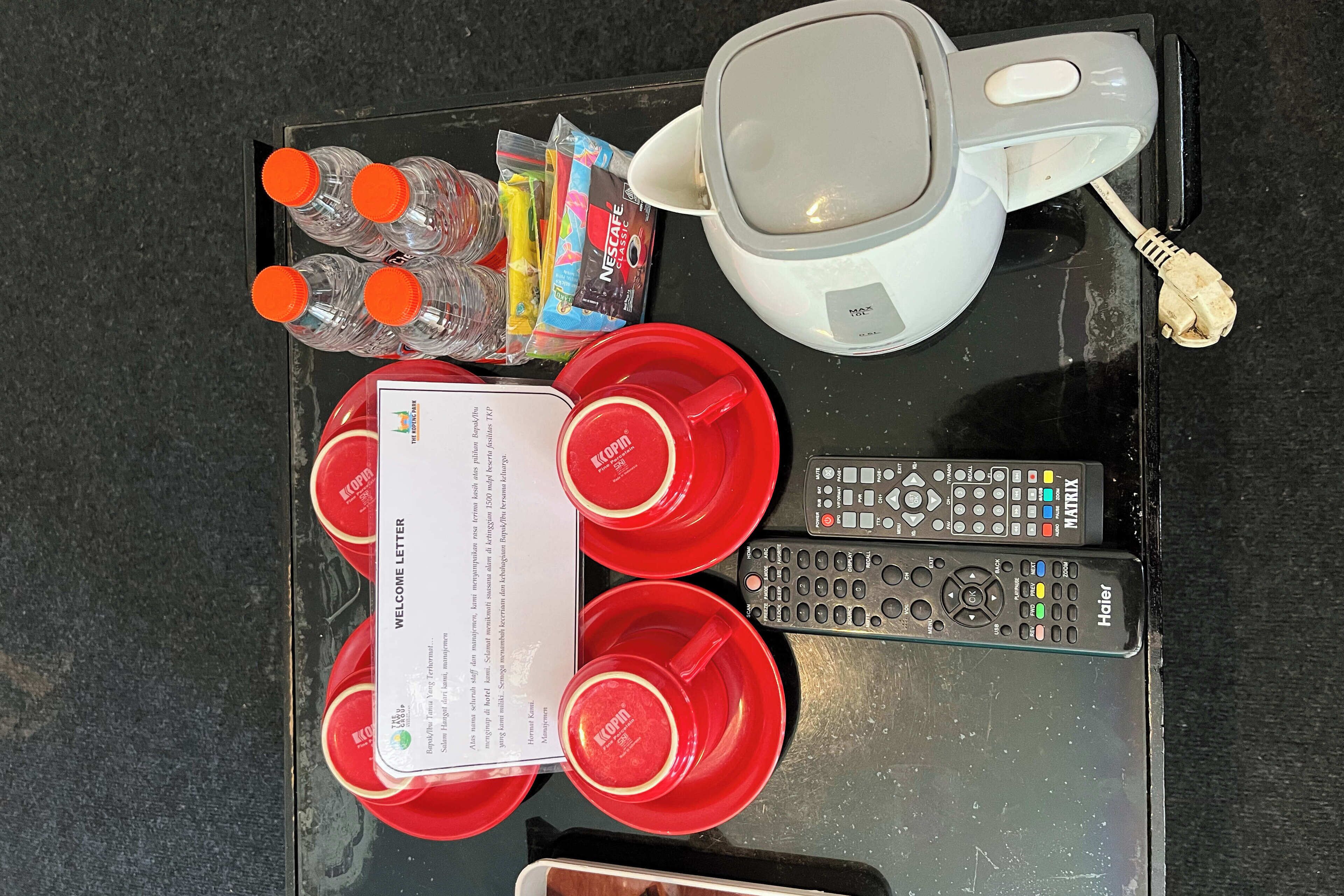 A tray with red cups, a white mug, bottled water, Nescafe packets, a welcome letter, and two remote controls.