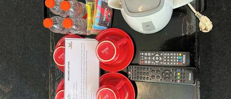 Uma bandeja com copos vermelhos, uma caneca branca, água engarrafada, sachês de Nescafé, uma carta de boas-vindas e dois controles remotos.