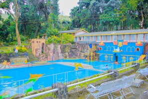 Una piscina con un mural al estilo de castillo, rodeada de follaje y un pequeño edificio con techo amarillo.
