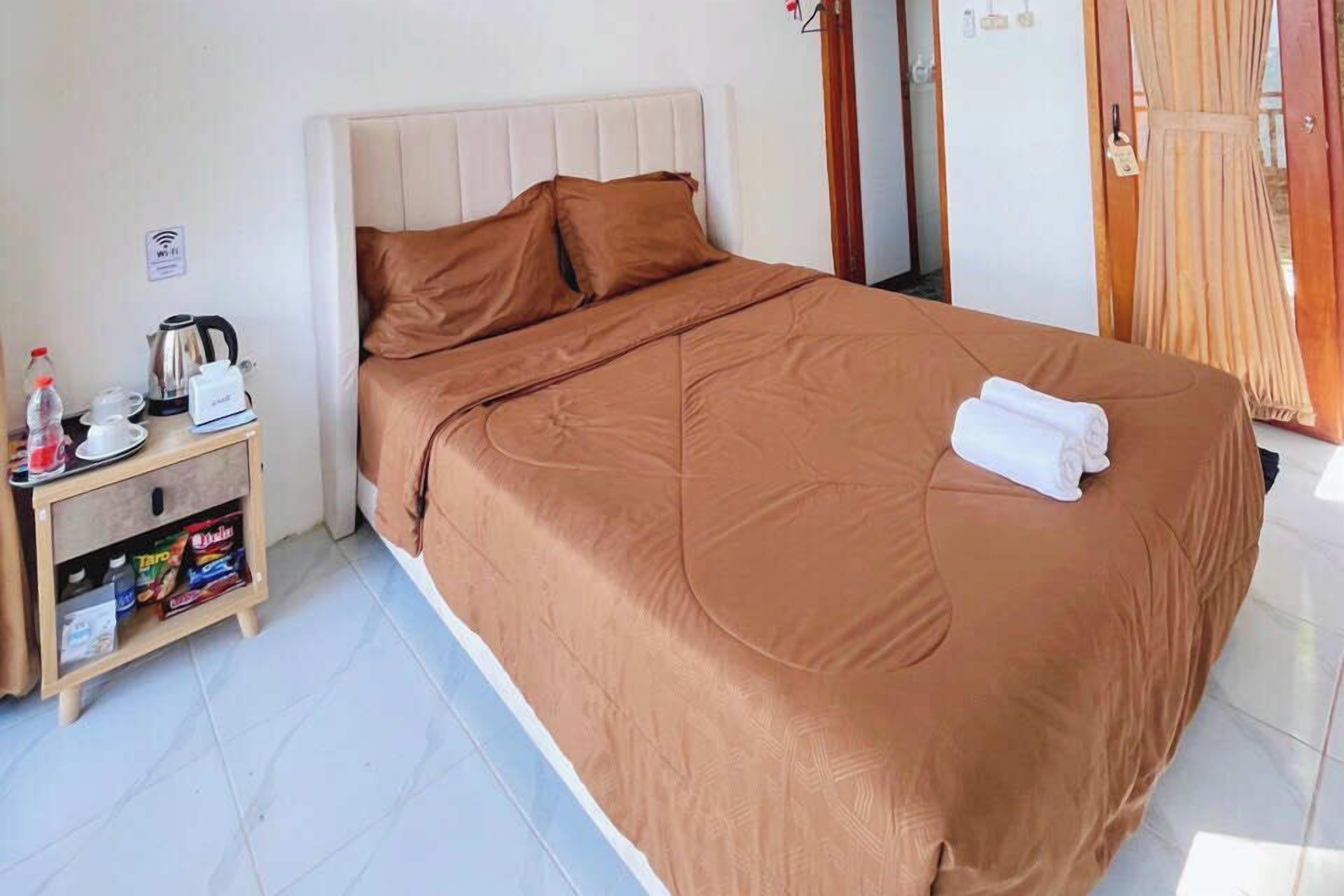 Una cama bien hecha con una colcha marrón y almohadas, una mesita de noche con una tetera, tazas y una caja de pañuelos.