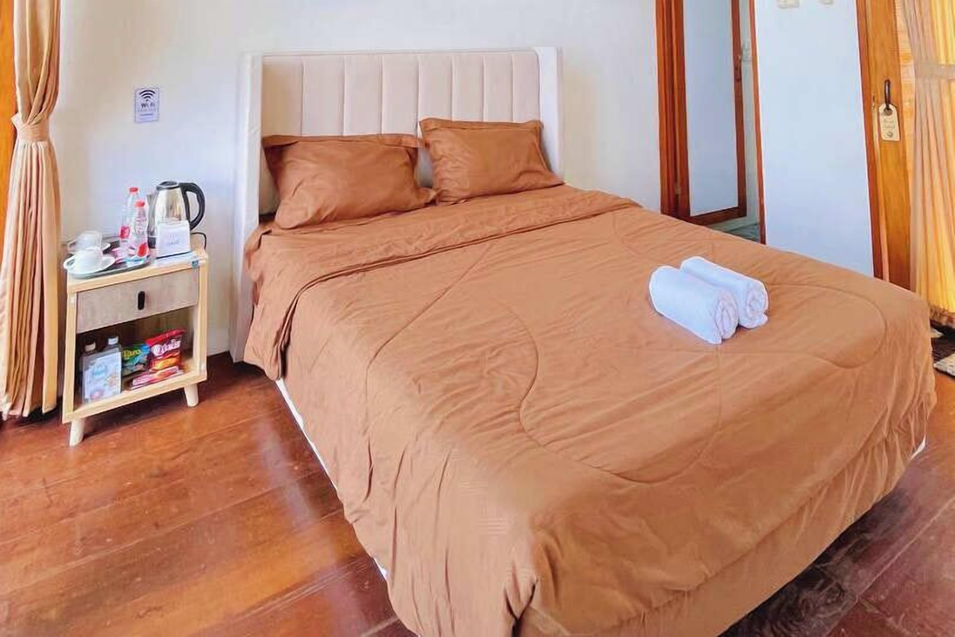 A neatly made bed with a matching headboard, a bedside table with a kettle and toiletries, and a wooden nightstand with a shelf.