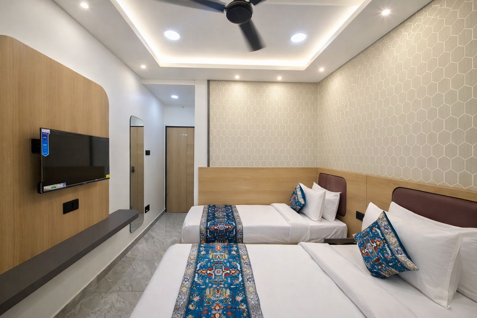 A hotel room with two beds, a flat-screen TV, a ceiling fan, and a hexagonal patterned wall.