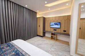 A modern bedroom with a bed, a TV mounted on a wooden panel wall, and a large window with curtains. - Matrushree Hotels - Near Shree Dwarkadhish Temple (Dwarka)